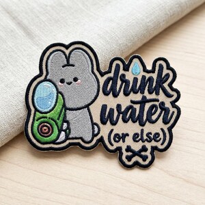 May include: An embroidered patch featuring a gray cartoon rabbit holding a green water gun with a blue water droplet. The patch includes the dark blue text "drink water (or else)" and a small black crossbones icon on a beige background.