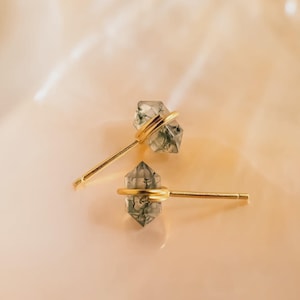 May include: A pair of stud earrings featuring clear, faceted crystals with green inclusions, each encircled by a gold-colored band. The earrings have gold-colored posts and are set against a soft, peach-colored background.