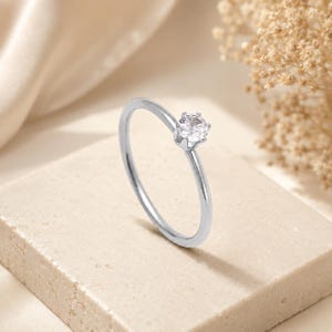 Può includere: Un anello sottile color argento con una singola pietra preziosa rotonda, trasparente e sfaccettata, montata su una montatura a sei griffe. La fascia è liscia e lucida, con un design essenziale che mette in risalto la luce della pietra.