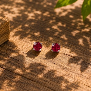 May include: A pair of stud earrings with vibrant red gemstones set in silver. The earrings are displayed on a wooden surface, with sunlight casting shadows. The gemstones are oval-shaped and appear to be rubies.