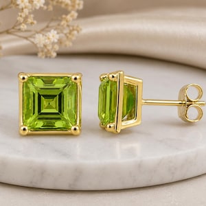 May include: A pair of stud earrings featuring square-cut, vibrant lime green gemstones set in polished yellow gold. Each earring has a four-prong setting and a standard post back, showcasing a faceted design that reflects light from the center.