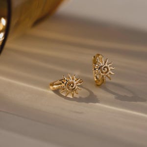 May include: A pair of gold hoop earrings featuring a sun-shaped charm with a central spiral and radiating rays. The earrings have a polished metallic finish and a hinged closure mechanism, capturing light to highlight the textured sun design.