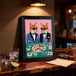 May include: Framed artwork featuring two foxes in suits and sunglasses, dining at a table with sushi and wine. The foxes are depicted on a pink background, with a green tablecloth. The artwork is in a black frame.
