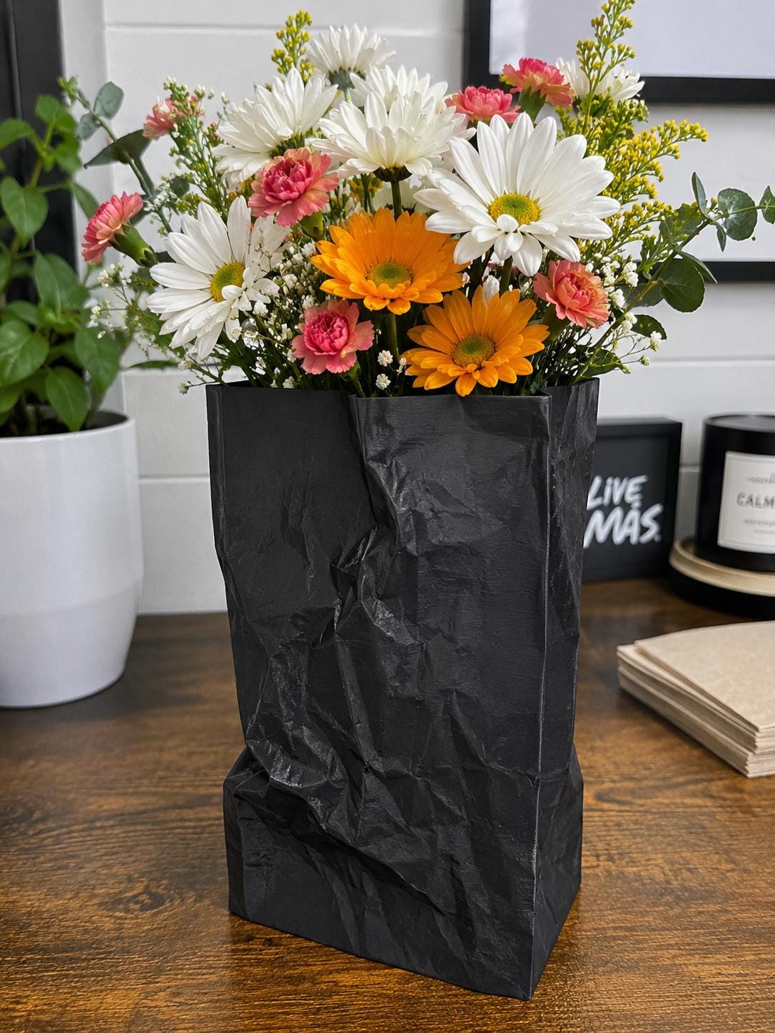 A black rigid container styled like a crumpled paper bag, filled with a vibrant bouquet of flowers.