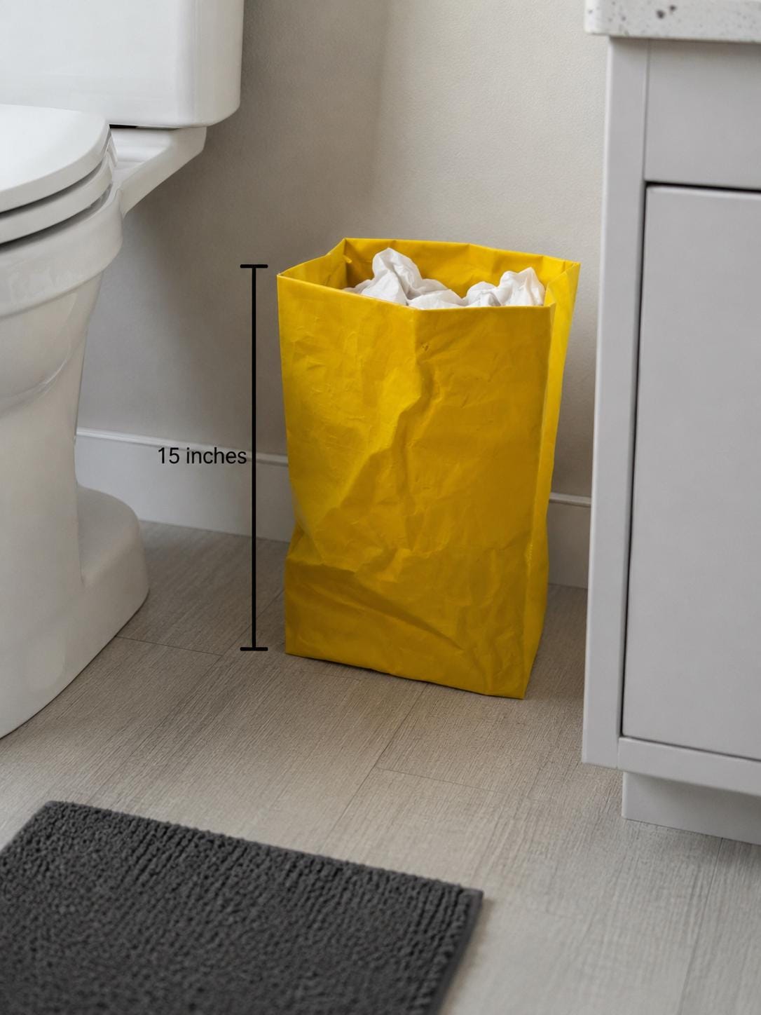 A 15-inch tall yellow paper bag replica used as a waste bin in a bathroom next to the toilet.