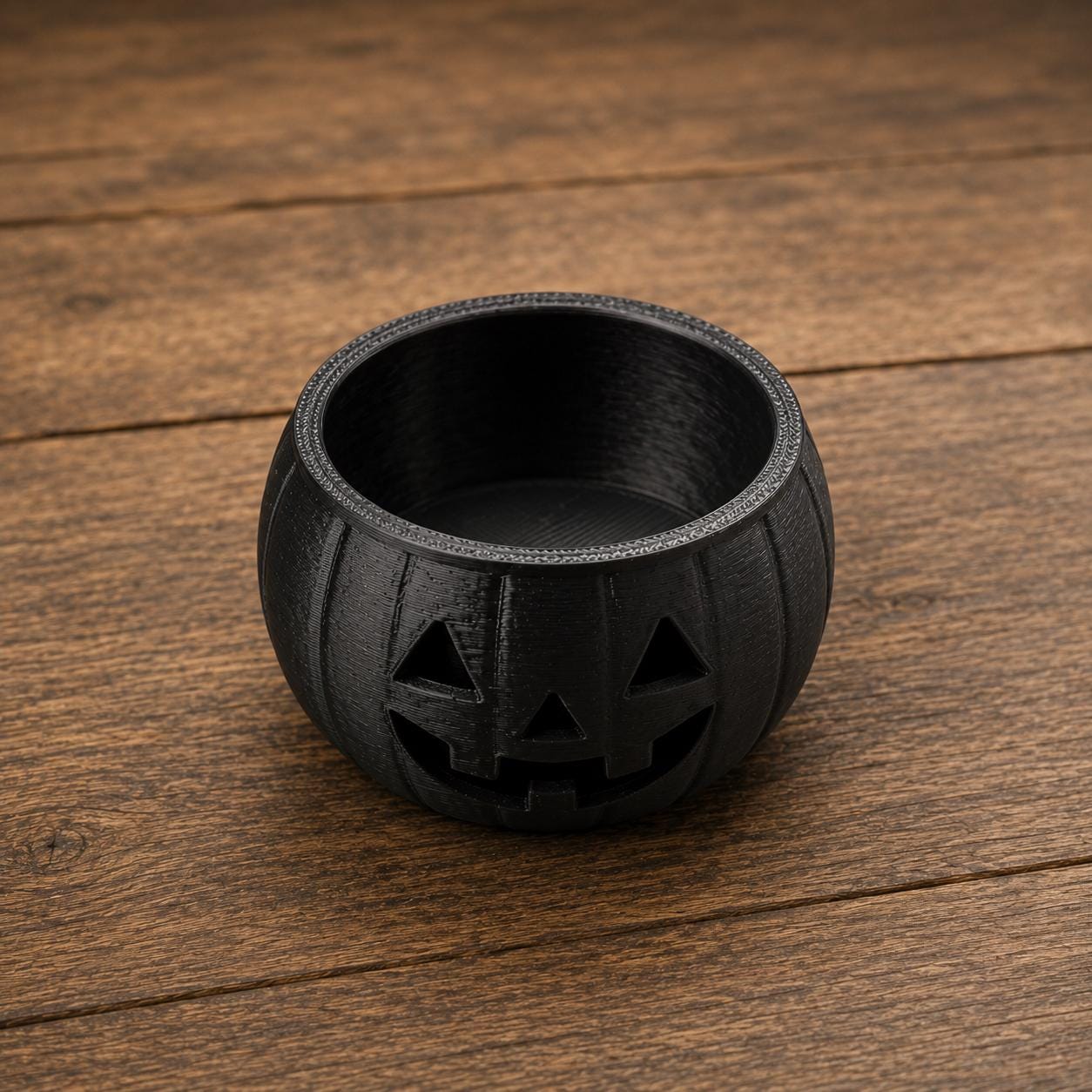 A small, black, 3D printed jack-o'-lantern candy bowl shown from a front-side angle on a wooden surface.