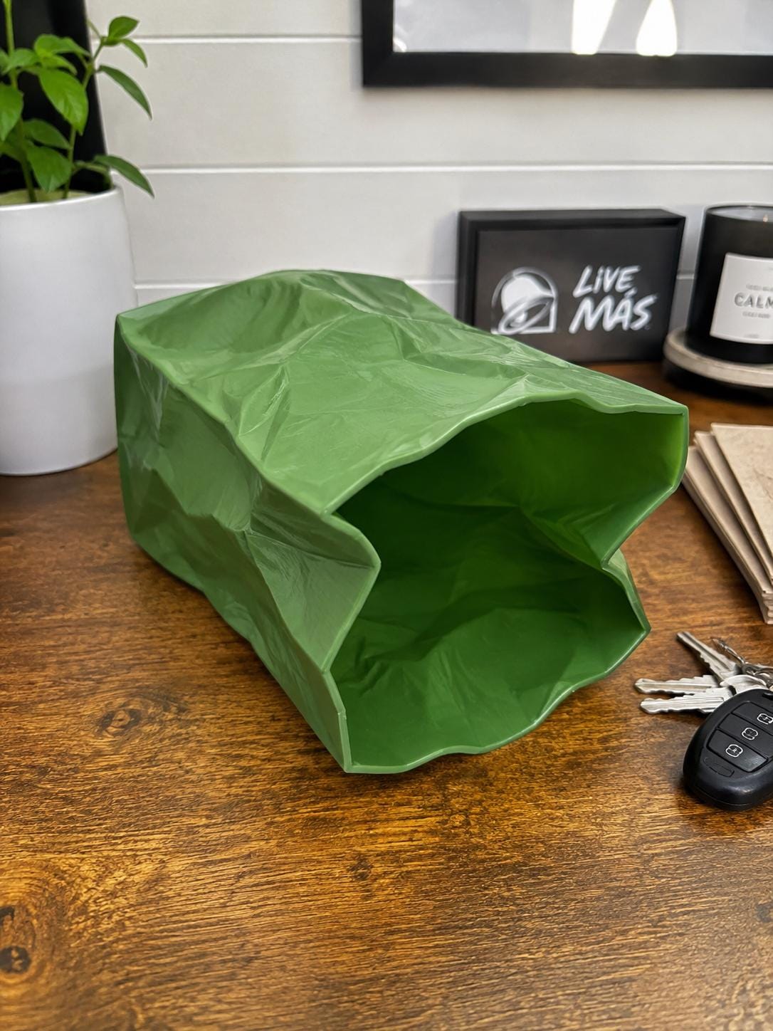 A green 3D printed paper bag replica sits on a wooden desk next to car keys and a plant.