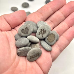 May include: A collection of smooth, gray river stones, some engraved with designs. One stone features a heart, another a pumpkin, and another has the name "Jesse". The stones are held in a hand.