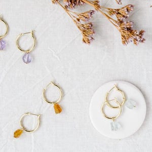 Puede incluir: Pendientes de aro dorados con colgantes de piedras preciosas en tonos morados, naranjas y azules. Los pendientes se exhiben sobre una superficie blanca con acentos florales secos, mostrando una colección de joyas hechas a mano.