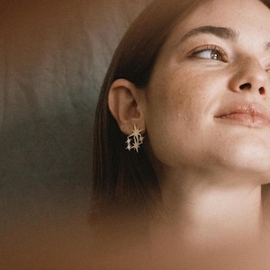 Könnte beinhalten: Eine Frau mit langen braunen Haaren trägt goldene sternförmige Ohrringe. Die Ohrringe hängen und haben ein geometrisches Design.