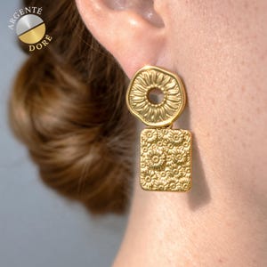 May include: Gold-toned earrings with a sunflower design. The top portion is a circular shape with a central hole, while the bottom is a rectangular shape. The earrings are embossed with a floral pattern. The text "ARGENTÉ DORÉ" is visible.