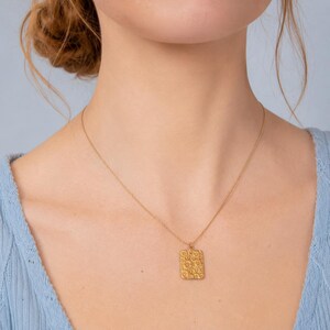 May include: A gold-colored necklace with a rectangular pendant. The pendant has a textured surface with a floral design. The necklace is worn on a person with a light blue top.