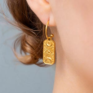 Peut inclure: Une boucle d'oreille créole dorée avec un pendentif rectangulaire. Le pendentif présente un motif en relief composé d'un œil central entouré de rayons, encadré par des croissants de lune et des étoiles sur les bords supérieur et inférieur.