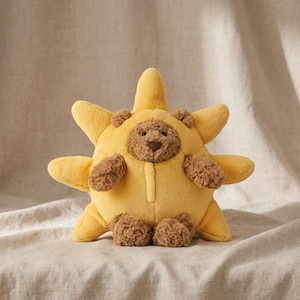 Puede incluir: Un juguete de peluche con forma de sol, con una cara de oso de peluche sonriente y patas marrones. El sol es amarillo con ocho puntas, y el oso tiene una nariz y orejas marrones. El juguete está hecho de material suave y afelpado.