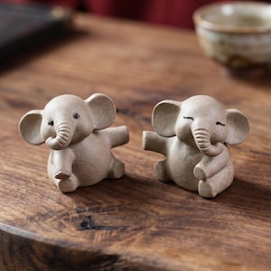 May include: Two small, beige ceramic elephant figurines with outstretched trunks and closed eyes. The elephants are on a wooden surface, with a small ceramic cup in the background. The figurines have detailed features and a whimsical design.