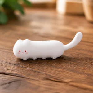 Puede incluir: Una pequeña figura blanca alargada con forma de gato y acabado mate liso. La figura presenta un rostro sencillo con dos pequeños puntos rojos como ojos, una boca roja diminuta y dos mejillas circulares de color rosa. Tiene una cola curva.