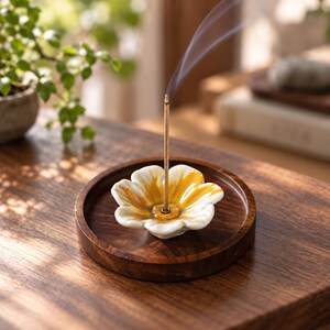 Könnte beinhalten: Ein Keramik-Räucherstäbchenhalter in Form einer weißen Blume mit gelben Akzenten, zentriert auf einem runden, dunkelbraunen Holztablett. Ein einzelnes dünnes Räucherstäbchen steht aufrecht in der Mitte der Blume und gibt grauen Rauch ab.