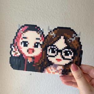 May include: Pixel art of two cartoon figures, one with pink hair and the other with brown hair and glasses. The figures are smiling and have small horns. The artwork is made of small, colorful beads.