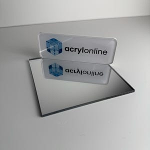 Puede incluir: Una placa de identificación rectangular de acrílico transparente con un logotipo de cubo azul y el texto negro acrylonline, colocada sobre una superficie de espejo cuadrada y reflectante que muestra un reflejo claro de la placa.