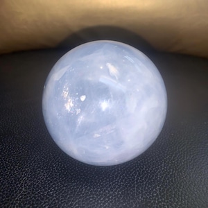 Könnte beinhalten: Eine hellblaue Kristallkugel. Die Kugel ist glatt und durchscheinend, mit subtilen Farbvariationen. Die Kristallkugel ist ein dekorativer Gegenstand, der oft zur Dekoration oder Meditation verwendet wird. Die Kugel liegt auf einer schwarzen Oberfläche.