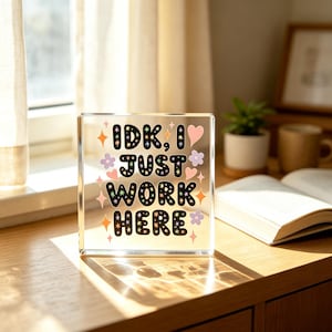 Könnte beinhalten: Ein durchsichtiger Acrylblock mit dem schwarzen Text IDK, I JUST WORK HERE in einer abgerundeten Schriftart. Das Design ist mit kleinen bunten Sternen, rosa Herzen und hellvioletten Blumen verziert, die um den Schriftzug auf der Oberfläche liegen.