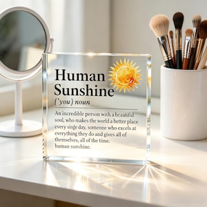 May include: A clear rectangular acrylic block featuring a printed definition of the term Human Sunshine. The text includes the word noun, a description of an incredible person, and a yellow sun illustration. The block has sharp edges and reflects light.