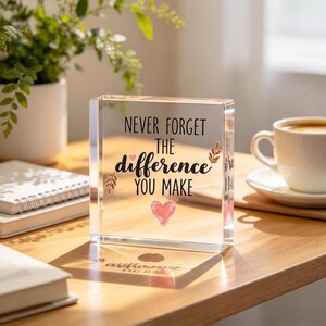 May include: A clear, rectangular acrylic block paperweight featuring the black printed text "NEVER FORGET THE DIFFERENCE YOU MAKE" with small decorative leaf illustrations and a pink heart graphic centred at the bottom of the block.