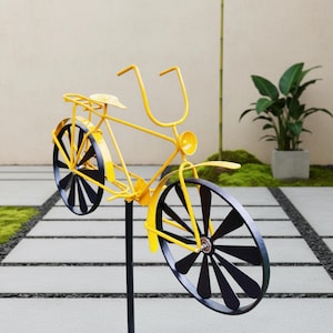 Puede incluir: Una pieza de arte de jardín con forma de bicicleta de metal amarillo con detalles en negro en las ruedas. La bicicleta decorativa tiene un asiento, manillar y faro. La bicicleta está montada en un poste negro y se exhibe al aire libre.