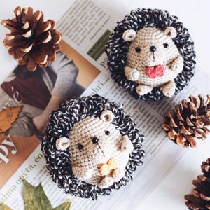 May include: Two handmade crochet hedgehog ornaments with black and white spines, tan faces, and small red and yellow accents. The hedgehogs are displayed on a newspaper with pinecones and fall leaves.