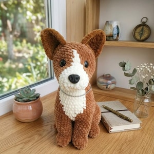 Puede incluir: Un perro de ganchillo marrón y blanco hecho a mano, con un pecho blanco y un collar de cadena plateada. El perro de peluche tiene ojos de botón negros y una nariz negra. Está sentado sobre una superficie de madera cerca de una ventana, con una pequeña planta suculenta en una maceta de terracota.