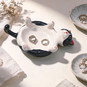 May include: A ceramic trinket dish shaped like a black and white cat lying on its back with its paws in the air. The cat has a white belly, black back, and a long tail. Two gold-toned rings are resting inside the bowl-shaped center of the dish.
