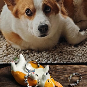 May include: A ceramic corgi-shaped ring holder in white, orange, and black, with two silver rings. The ring holder is lying on its back on a wooden surface. A real corgi dog is in the background.