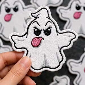 May include: A white embroidered ghost patch with black outlines, featuring an angry facial expression and a pink tongue sticking out. The ghost has a rounded head, two arms extended to the sides, and a draped body shape with stitched details.