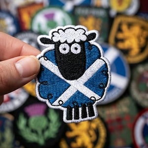 May include: An embroidered patch shaped like a sheep with a black face and white wool top. The sheep's body features a blue background with a white diagonal cross pattern, resembling the Scottish flag. The patch has a white stitched border.