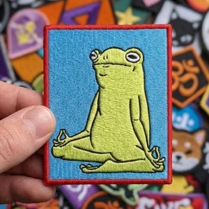 May include: A rectangular patch featuring a yellow frog in a meditative pose against a light blue background with a red border. The patch is held by a hand, surrounded by other colorful patches with various designs, including an Om symbol and a Shiba Inu.