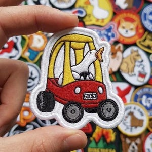 May include: Embroidered patch featuring a white goose in a red and yellow car with the text "GOOS3". The patch is surrounded by a white border and set against a background of other animal-themed patches.