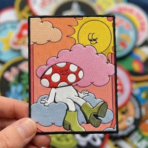 May include: A rectangular embroidered patch featuring a cartoon mushroom character with a red and white spotted cap, wearing green boots, sitting on a blue cloud. The background includes a pink cloud, a yellow sun, and an orange sky.