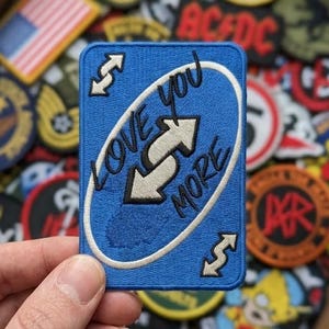 May include: A blue patch with the text "LOVE YOU MORE" and an arrow symbol. The patch is surrounded by other patches with various designs, including an American flag and the AC/DC logo.