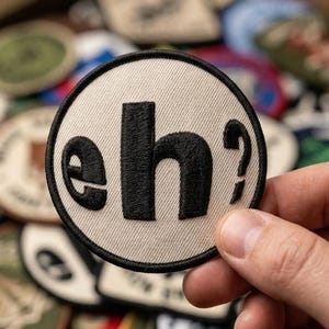 May include: A round, embroidered patch with the word "eh?" in black letters on a cream-colored background. The patch is held in a hand, with other patches visible in the background. The patch has a black border.