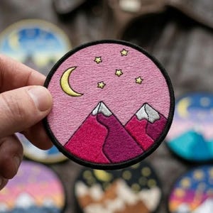 May include: A circular embroidered patch featuring a pink sky with a yellow crescent moon and small yellow stars above two mountain peaks. The mountains are rendered in shades of magenta and purple with white snow-capped tops and a black border.