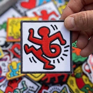 May include: A square patch with a red figure in motion, a design by Keith Haring, on a white background. The patch is held in a hand, with other colorful patches visible in the background. The patches are likely for clothing or accessories.