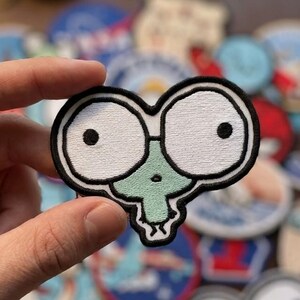 May include: Embroidered patch featuring a cartoon character with large, white eyes and a green face. The patch has a black border and is held in a hand. Other patches are in the background.