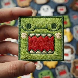 May include: A square embroidered patch featuring a green character with a wide, open mouth showing sharp white teeth against a red background. The character has two dark green circular eyes and small white flower-like accents on its cheeks.
