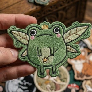 May include: Embroidered green frog patch with fairy wings, a gold crown, and a star pendant. The frog has large black eyes and pink cheeks. The patch is held in a hand, with other patches in the background.