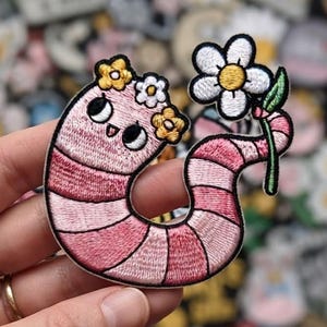 May include: An embroidered patch featuring a pink and light pink striped worm with a smiling face, large black eyes, and a crown of three small yellow and white flowers. The worm is holding a single white daisy with a yellow center and green stem.