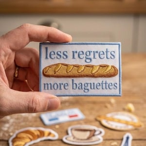 May include: A rectangular patch with the text "less regrets more baguettes" above an embroidered baguette. Other patches include a croissant, coffee cup, Eiffel Tower, and wheat design.
