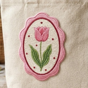 May include: A beige canvas tote bag with a pink embroidered oval patch. The patch features a pink tulip with green leaves and red dots, framed by a scalloped pink border. The bag has two beige handles.
