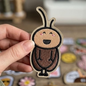 May include: An embroidered patch featuring a cartoon character with a tan oval head, pink cheeks, a wide black smile, and two antennae. The character has a dark brown rounded body with thin black arms and legs, all outlined in black thread.