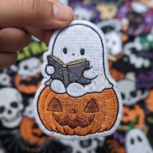 May include: An embroidered patch featuring a white ghost with a small smile reading a black book while sitting atop a textured orange jack-o'-lantern with a carved face, finished with a white stitched border against a dark green background.