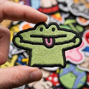 May include: A green frog patch with a black outline and a pink tongue. The patch is held in a hand, with other colorful patches in the background. The frog patch is approximately 2 inches wide.